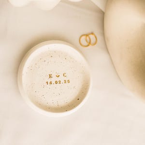 May include: A small, round, speckled, off-white ceramic ring dish with gold lettering. The dish has the initials "E C" with a heart symbol and the date "14.02.25". Two gold rings are next to the dish. The background is a white cloth.