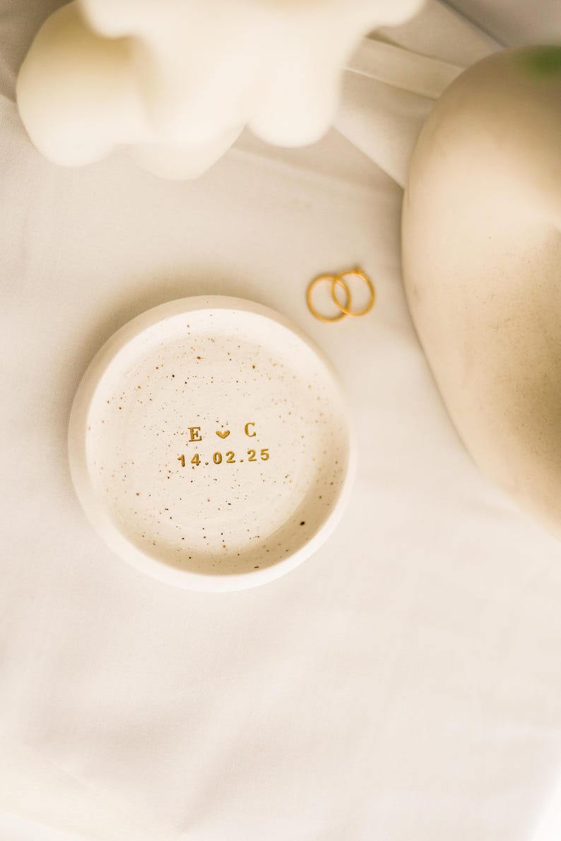 May include: A small, round, speckled, off-white ceramic ring dish with gold lettering. The dish has the initials "E C" with a heart symbol and the date "14.02.25". Two gold rings are next to the dish. The background is a white cloth.