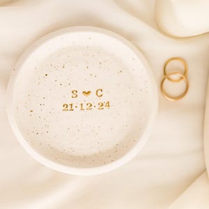 May include: A speckled, off-white ceramic ring dish with gold lettering and a heart symbol. The dish displays the initials "S C" and the date "21.12.24". Two gold rings are next to the dish on a cream-colored fabric.