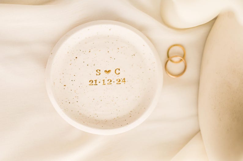May include: A speckled, off-white ceramic ring dish with gold lettering and a heart symbol. The dish displays the initials "S C" and the date "21.12.24". Two gold rings are next to the dish on a cream-colored fabric.