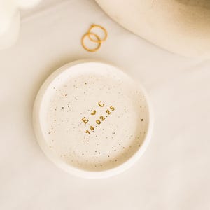 May include: A speckled, off-white ceramic ring dish with gold lettering. The dish has the initials "E C", a heart symbol, and the date "14.02.25". Two gold rings are next to the dish. The background is a soft, neutral colour.
