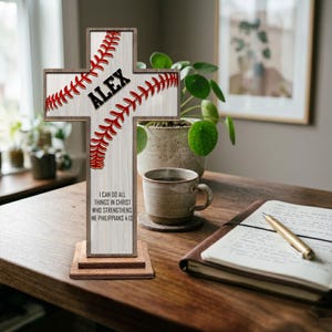 Peut inclure: Une croix en bois sur le thème du baseball. La croix présente des coutures de baseball rouges et le nom "ALEX" en noir. La base porte l'inscription "I CAN DO ALL THINGS IN CHRIST WHO STRENGTHENS ME PHILIPPIANS 4:13".
