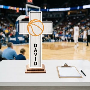 Peut inclure: Une croix en bois avec un motif de ballon de basket et le nom "DAVID" en lettres noires. La croix est blanche avec une base en bois marron. Un ballon de basket orange est centré sur la croix. Un presse-papiers et un stylo sont sur la table.