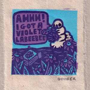 以下が含まれることがあります： 「AHHH！I GOT A VIOLET LABEEBEE」という吹き出しを持つ生き物を描いた、気まぐれでカラフルなプリント。アートワークは紫、青、白の色合いを使用し、花のモチーフとアーティストのサイン「GOOBER」が特徴です。