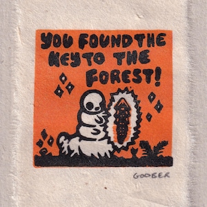 以下が含まれることがあります： 「YOU FOUND THE KEY TO THE FOREST!」の文字と、毛虫のような漫画のキャラクターが描かれたオレンジ色の正方形のアートプリント。 イラストは白黒で、背景は白です。