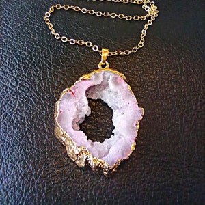 May include: A necklace featuring a gold-tone chain and a pendant made from a slice of geode with a light pink, crystalline center. The irregular, organic-shaped stone is framed by a metallic gold-colored edge, creating a distinct contrast.