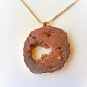 May include: A necklace featuring a circular, brown, geode-style pendant with a hollow centre and crystalline texture. The pendant is edged in a metallic copper tone and suspended from a delicate, matching gold-coloured metal chain link necklace.