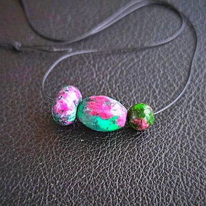 May include: Three natural gemstone beads on a black string. The beads are a mix of colors, including pink, green, and white. The beads are of varying shapes and sizes, with a polished finish. The background is a textured, dark surface.