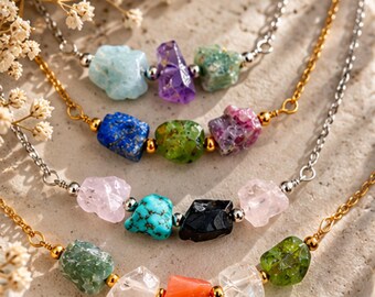 May include: Four necklaces featuring raw, irregularly shaped gemstones in various colors including blue, purple, green, pink, turquoise, black, orange, and clear. The stones are set on thin gold and silver-toned metal chains with small metallic beads.