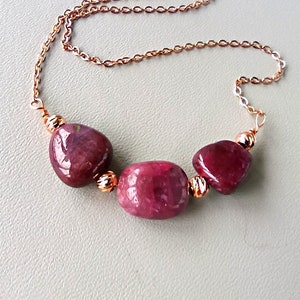 Könnte beinhalten: Halskette aus Roségold mit drei polierten, tiefroten Edelsteinen. Die unregelmäßig geformten Edelsteine sind an einer filigranen Kette mit dekorativen goldfarbenen Perlen aufgereiht. Die Halskette wird auf einer hellgrauen Oberfläche präsentiert.