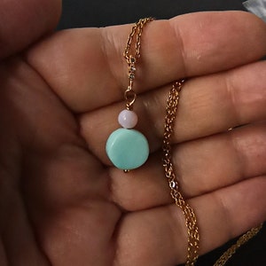 May include: A gold-colored chain necklace with a pendant featuring a light blue, round stone and a smaller pink stone. The pendant is connected to the chain with a small, sparkling accent. The necklace is displayed against a dark background.