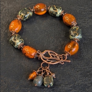 Könnte beinhalten: Ein Armband mit abwechselnd glatten orangefarbenen und gesprenkelten grünen Perlen, die durch kupferfarbene Metallabstandshalter getrennt sind. Das Schmuckstück hat einen blattförmigen Knebelverschluss aus kupferfarbenem Metall mit drei kleinen Anhängern.