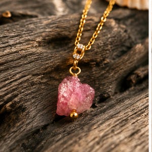 Può includere: Una collana a catena dorata con un ciondolo in pietra grezza rosa a forma di cuore. Il ciondolo è impreziosito da una piccola perlina dorata sul fondo e da una contromaglia dorata incastonata con due minuscole pietre chiare.