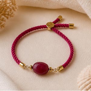 Op de afbeelding: Een magenta koordarmband met een centrale gefacetteerde donkerrode ovale edelsteen. De armband is voorzien van goudkleurige metalen accenten, waaronder twee kleine kraaltjes naast de steen, twee eindkapjes en een verstelbare schuifsluiting met bedel.