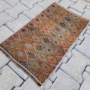 May include: A small, rectangular rug with a geometric pattern in shades of brown, orange, and cream. The rug has a fringe border.