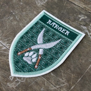 Op de afbeelding: Geborduurde patch in schildvorm, met het woord "RANGER" in het wit. De patch heeft een groene achtergrond met een patroon, gekruiste messen en een pootafdruk. De rand is lichtgroen.