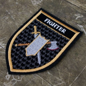 May include: Embroidered patch with a black shield shape, featuring a sword, shield, and axe design. The word "FIGHTER" is embroidered in white on a black banner at the top. The patch has a gold border and is set against a textured background.