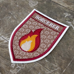 Op de afbeelding: Geborduurde patch in schildvorm, met het woord "SORCERER" in wit op een bruine banner. De patch heeft een rode rand en een vlamontwerp in rood, geel en wit. De achtergrond is bruin met een sterrenpatroon.