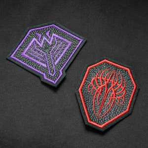 Könnte beinhalten: Zwei gestickte Aufnäher. Einer ist eine lila und schwarze Raute mit einem geometrischen Design. Der andere ist ein rotes und schwarzes Achteck mit einem stilisierten Design. Beide Aufnäher haben schwarze Ränder auf schwarzem Hintergrund.