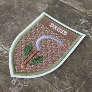 May include: Embroidered patch with a shield shape, featuring the word "DRUID" in white letters on a brown banner. The patch has a brown background with a repeating pattern, a silver sickle, and green leaves. The patch is bordered with green and white.