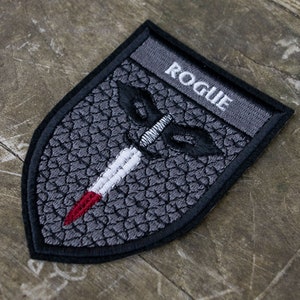 May include: Embroidered patch in a shield shape with the word "ROGUE" at the top. The patch features a dagger design with a white blade and red tip, set against a textured gray background. The patch is outlined in black.