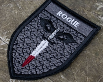 May include: Embroidered patch in a shield shape with the word "ROGUE" at the top. The patch features a dagger design with a white blade and red tip, set against a textured gray background. The patch is outlined in black.
