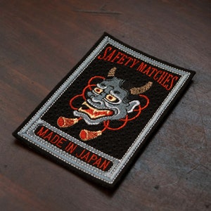 May include: Embroidered patch with a black background, featuring a gray demon face with red and brown accents. The text "SAFETY MATCHES" and "MADE IN JAPAN" are in red. The patch has a gray border.