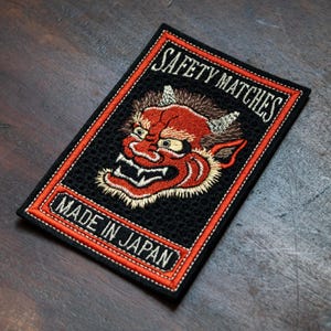 May include: Embroidered patch with a red demon face design, black background, and the words "SAFETY MATCHES" and "MADE IN JAPAN" in white. The patch has a red and orange border.