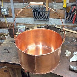 May include: A polished copper bucket with a cylindrical shape and a smooth, reflective surface. The bucket features a thin, arched metal handle attached to two side brackets, with a light-colored wooden grip centered on the top of the handle.