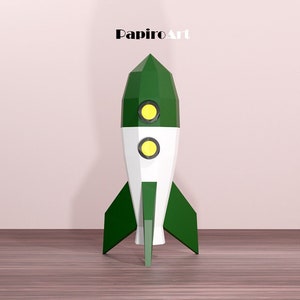 May include: A green, white, and yellow rocket ship model with the text "PapiroArt" at the top. The rocket has a pointed top, two yellow circular windows, and green fins. It is set against a light pink wall and a wooden floor.