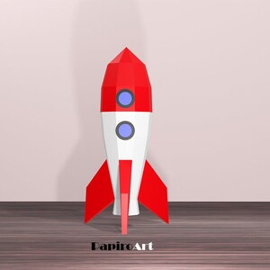 May include: A red, white, and blue geometric rocket model. The rocket has a pointed red top, a white midsection with two blue circular windows, and red fins. The rocket is standing on a wooden surface.