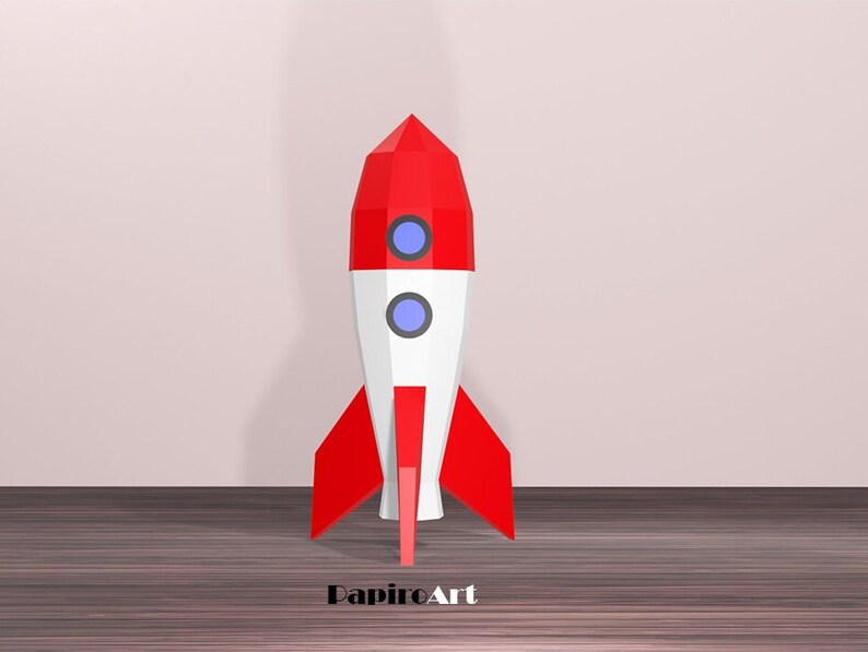 May include: A red, white, and blue geometric rocket model. The rocket has a pointed red top, a white midsection with two blue circular windows, and red fins. The rocket is standing on a wooden surface.