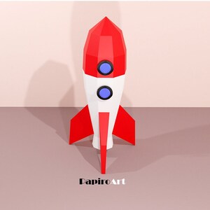 May include: A geometric rocket model in red, white, and blue. The rocket features a pointed top, two circular windows, and red fins. The word "PapiroArt" is printed at the bottom.