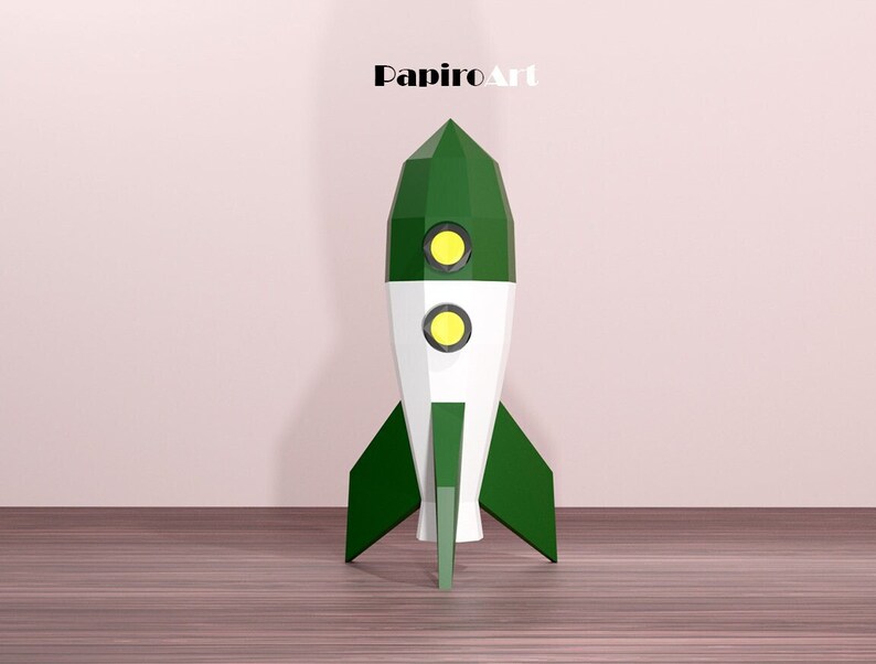 May include: A green, white, and yellow rocket ship model with the text "PapiroArt" at the top. The rocket has a pointed top, two yellow circular windows, and green fins. It is set against a light pink wall and a wooden floor.