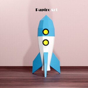 May include: A geometric rocket ship in blue, white, and yellow. The rocket has a pointed top, two circular windows, and blue fins. The text "PapiroArt" is at the top.