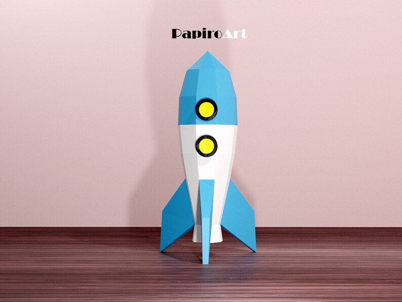 May include: A geometric rocket ship in blue, white, and yellow. The rocket has a pointed top, two circular windows, and blue fins. The text "PapiroArt" is at the top.