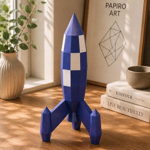 May include: A geometric rocket sculpture featuring a faceted design with a blue and white checkered pattern. The rocket stands on three pointed fins and has a conical top, constructed with sharp, angular edges that create a modern, structured appearance.