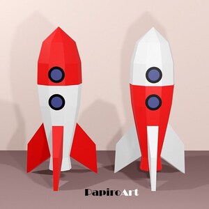 May include: Two papercraft rockets, one red and white, the other white and red, stand side-by-side. Each rocket has two blue circular windows and red fins. The rockets are set against a neutral background, with the text "PapiroArt" in the lower right corner.