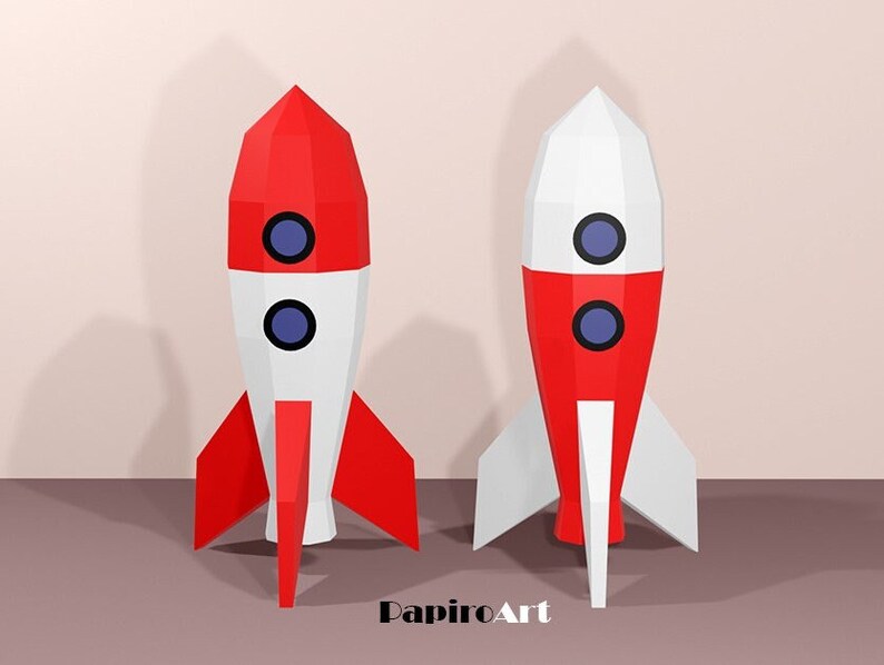 May include: Two papercraft rockets, one red and white, the other white and red, stand side-by-side. Each rocket has two blue circular windows and red fins. The rockets are set against a neutral background, with the text "PapiroArt" in the lower right corner.