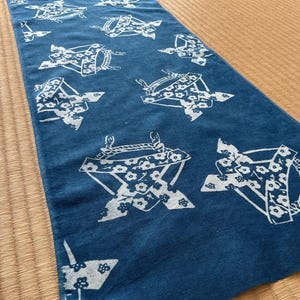 Könnte beinhalten: Ein blauer Stoffläufer mit einem sich wiederholenden weißen Blumen- und geometrischen Muster. Das Design zeigt stilisierte Formen, die an traditionelle japanische Gegenstände erinnern. Der Läufer wird auf einer gewebten Matte präsentiert.