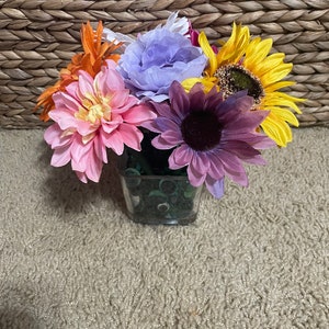 May include: A bouquet of artificial flowers in a clear glass vase. The flowers include a pink dahlia, a purple daisy, a yellow sunflower, and a light purple rose. The vase is filled with green beads.