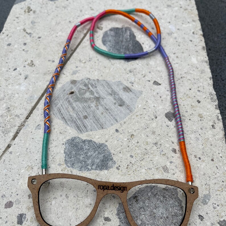 May include: A pair of wooden-framed glasses with a colorful, striped eyeglass chain. The chain is made of orange, pink, blue, and silver thread. The glasses have the words "ropa.design" printed on the frame.