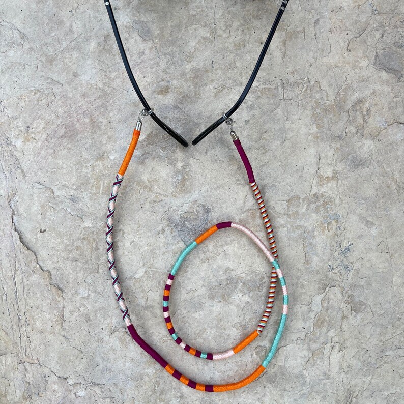 May include: A pair of glasses with a colorful, braided cord. The cord is made of orange, pink, blue, and white thread. The cord is attached to the glasses with metal clasps.