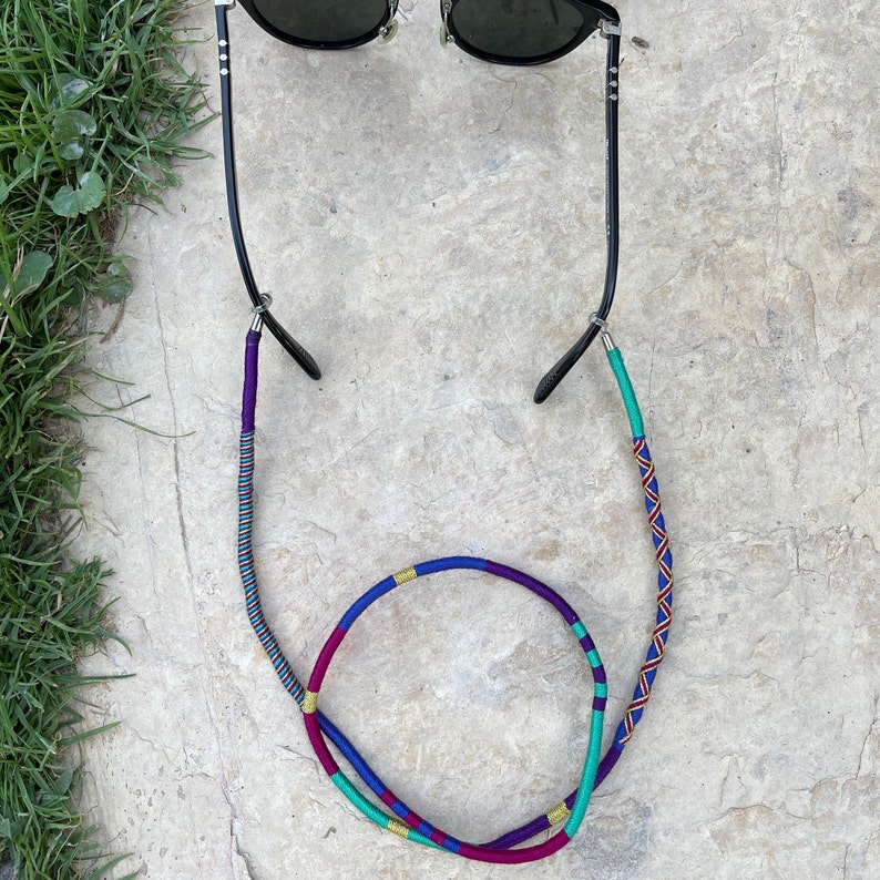 May include: A pair of black sunglasses with a colorful, braided cord. The cord is made of purple, blue, green, and gold thread.