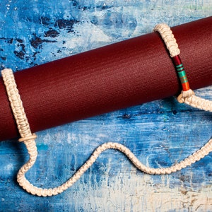 May include: A maroon yoga mat with a white braided strap. The strap has a colorful accent in the middle.