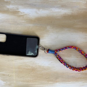 May include: A black phone case with a clear window on the back is attached to a braided phone strap. The strap is made of orange, pink, and blue cord.