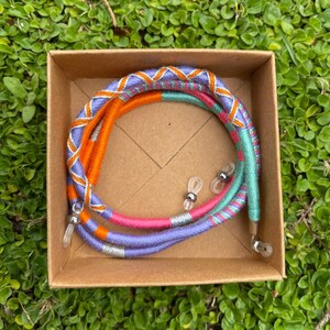 May include: A set of colorful glasses retainers in a cardboard box. The retainers are made of woven fabric in shades of orange, purple, pink, and green. The retainers have silver metal ends.