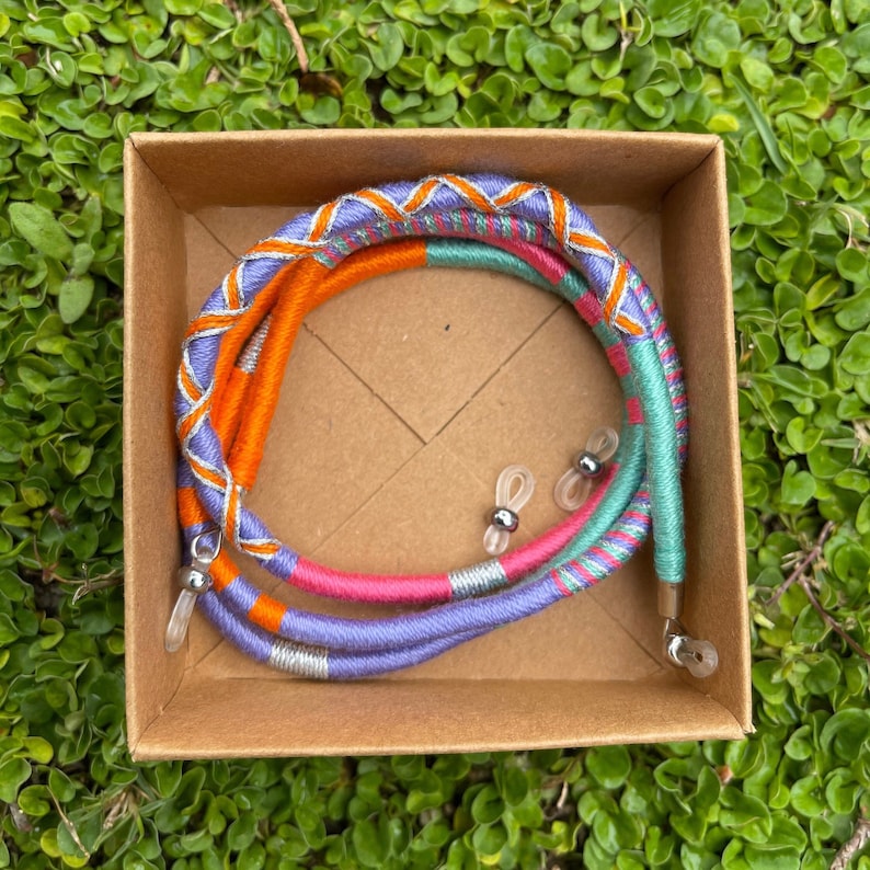 May include: A set of colorful glasses retainers in a cardboard box. The retainers are made of woven fabric in shades of orange, purple, pink, and green. The retainers have silver metal ends.