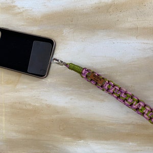 May include: A green and purple braided phone strap with a brown leather tag that says "ROPA" on it. The strap is attached to a black phone case.