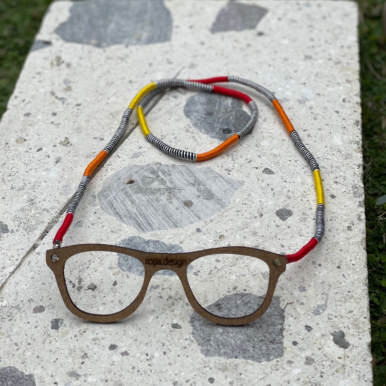 May include: A pair of wooden eyeglasses with a red, yellow, and black patterned eyeglass chain. The chain is made of woven string and has the text "ropa design" printed on the glasses.